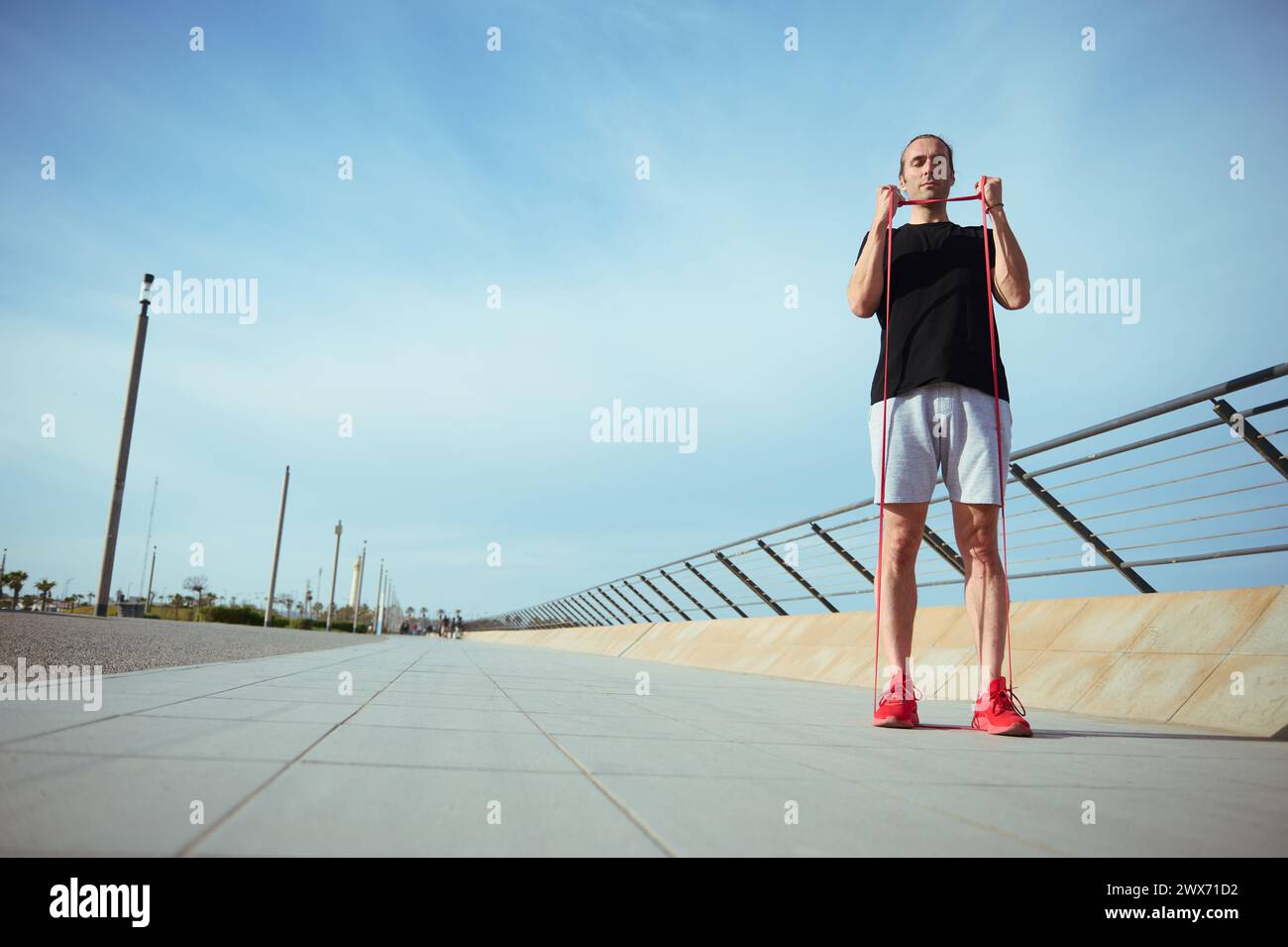 Man with athletic body performs bodyweight fitness exercises using a resistance band. Muscular guy in black t-shirt, gray sports shorts and red sneake Stock Photo