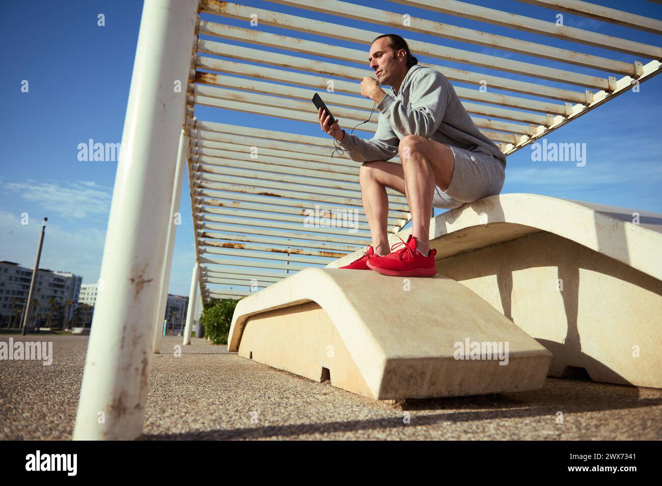 Full length portrait of Caucasian young handsome sporty man using smart mobile phone while having exercise break in the outdoors urban sports ground, Stock Photo