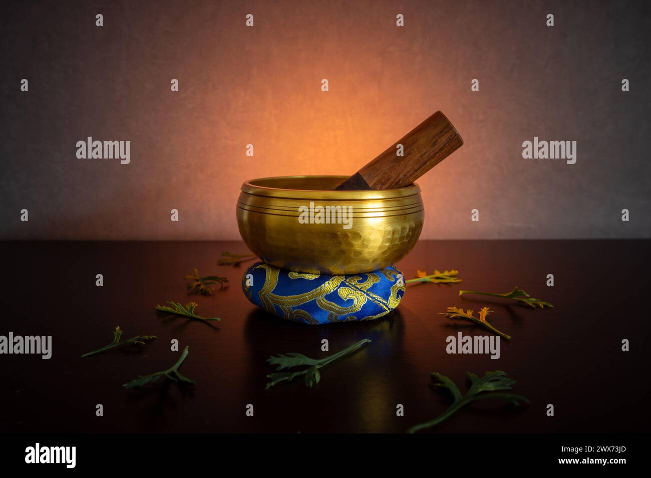 A Tibetan singing bowl sits on a ring cushion amid parts of leaves and flowers, bathed in golden backlight against a dark background Stock Photo