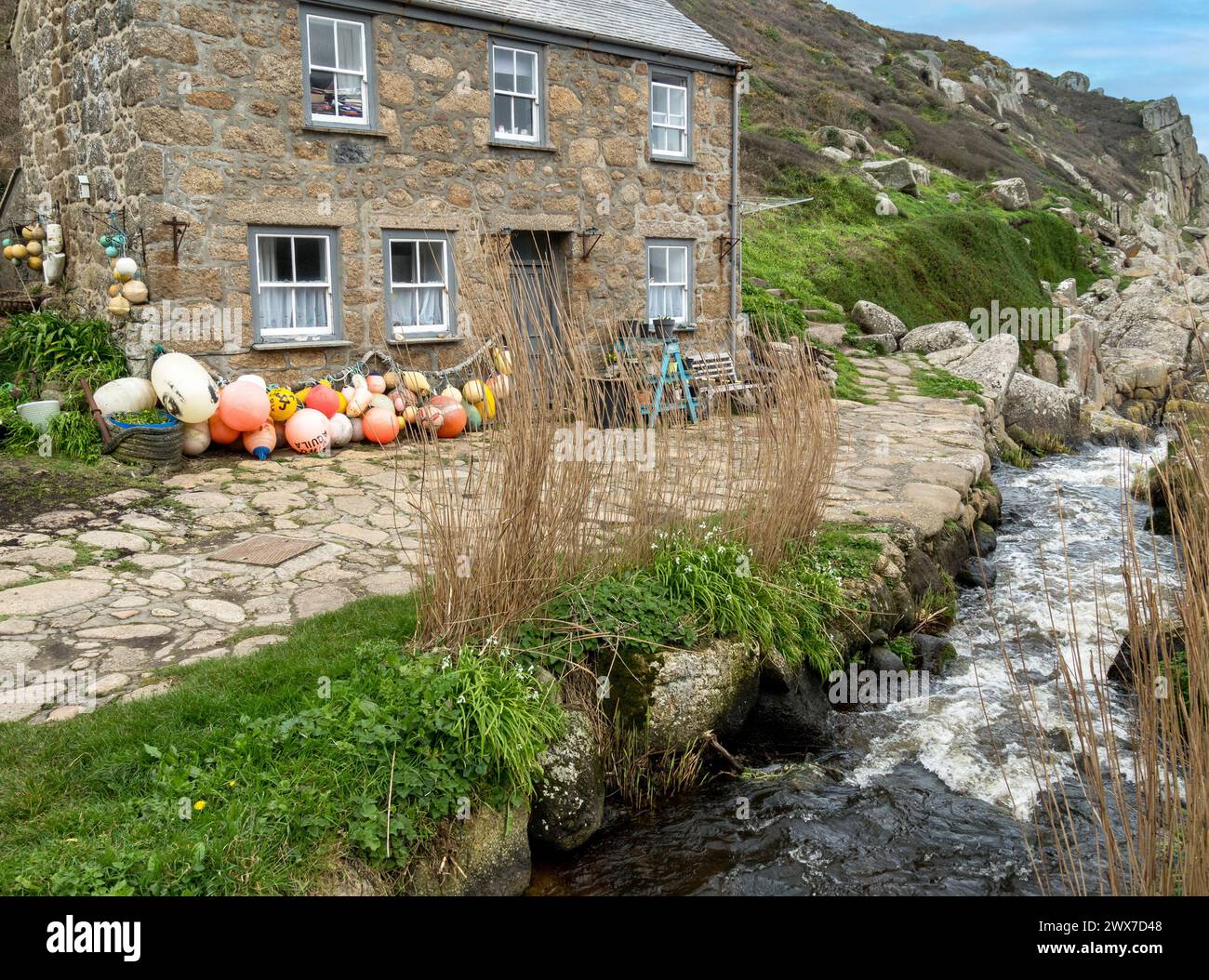 Attractive old granite stone Cornish seaside cottage by stream with fishing floats and buoys at Penberth Cove, Cornwall, UK Stock Photo