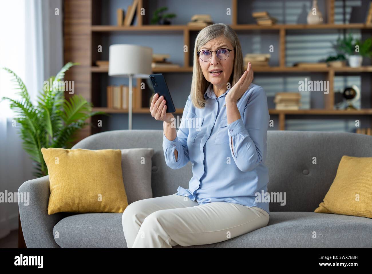 A mature adult woman sitting on a couch appears shocked and in disbelief while on a phone call in a cozy home setting. Stock Photo