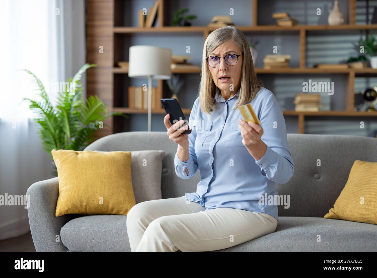 A mature adult woman examines her credit card while holding a smartphone, sitting comfortably on her couch at home, possibly suspicious of fraud. Stock Photo