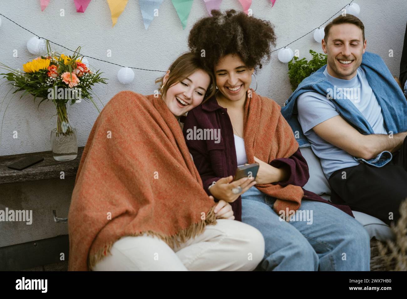 Happy female friends wrapped in blanket and sharing smart phone at party with friends in balcony Stock Photo
