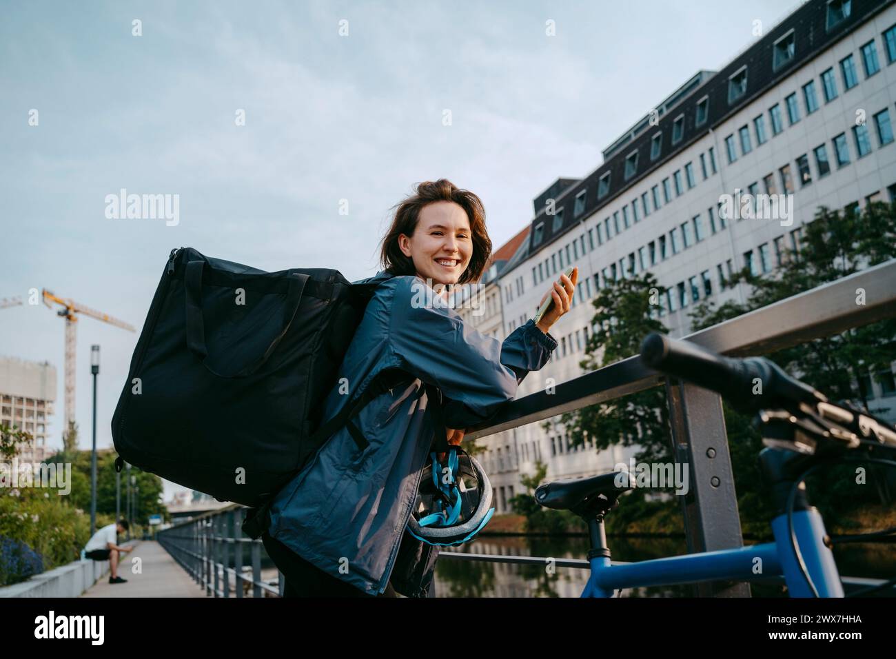Side view portrait of smiling female delivery person holding smart phone while leaning on railing in city Stock Photo