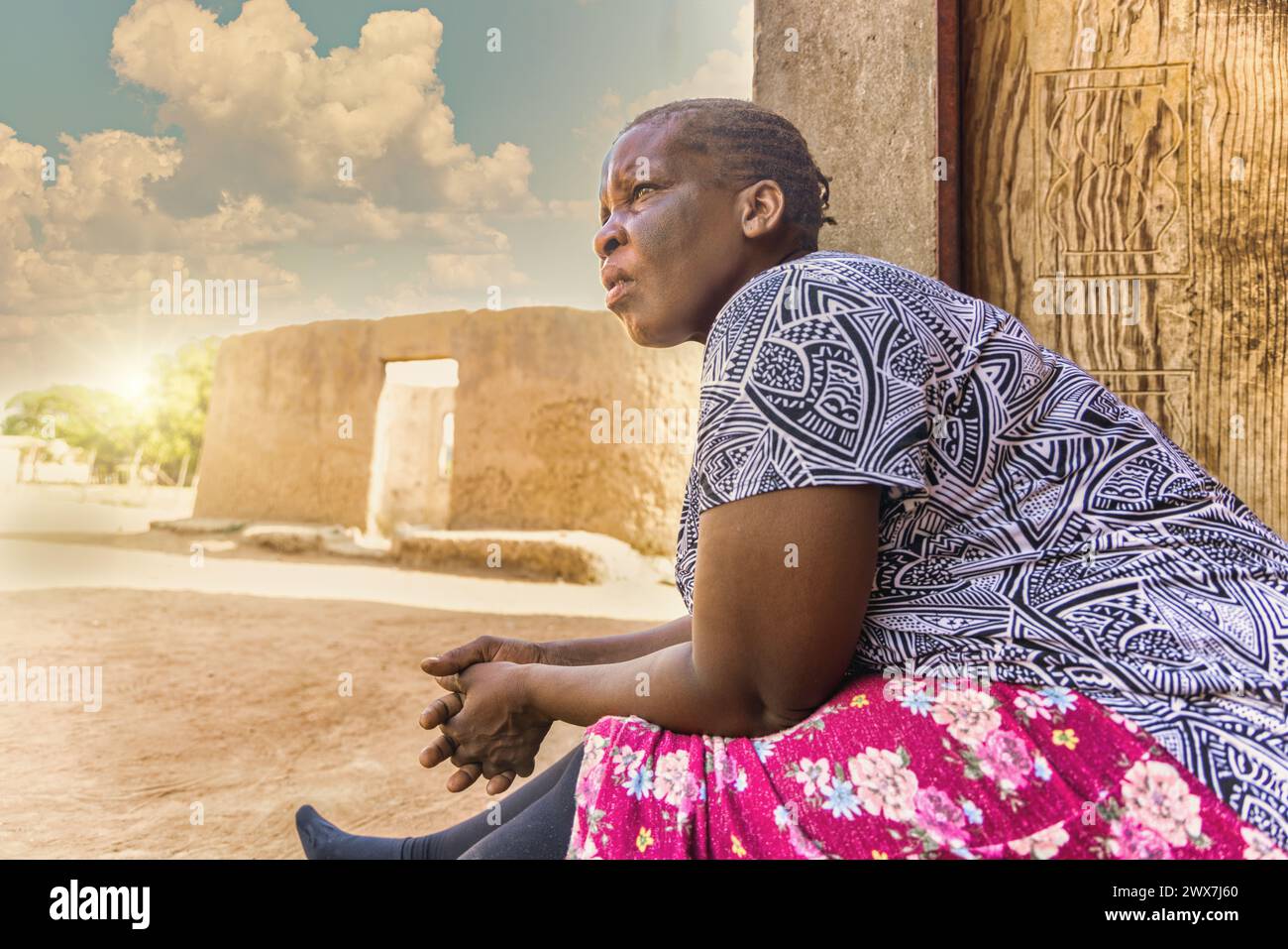 old african woman sitting on the veranda, ruined mud house in the background, african village life Stock Photo
