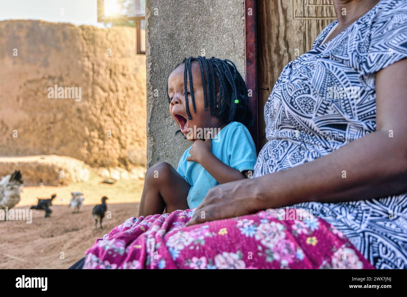 sleepy tired african child with braids sitting together with her granny on the veranda, chickens running, , african village life Stock Photo