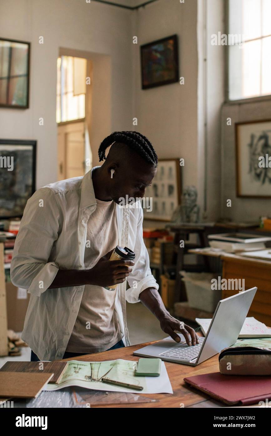 Male entrepreneur with disposable cup using laptop while standing at workshop Stock Photo