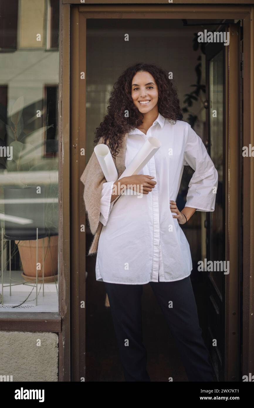 Portrait of smiling female architect standing with hand on hips at office doorway Stock Photo