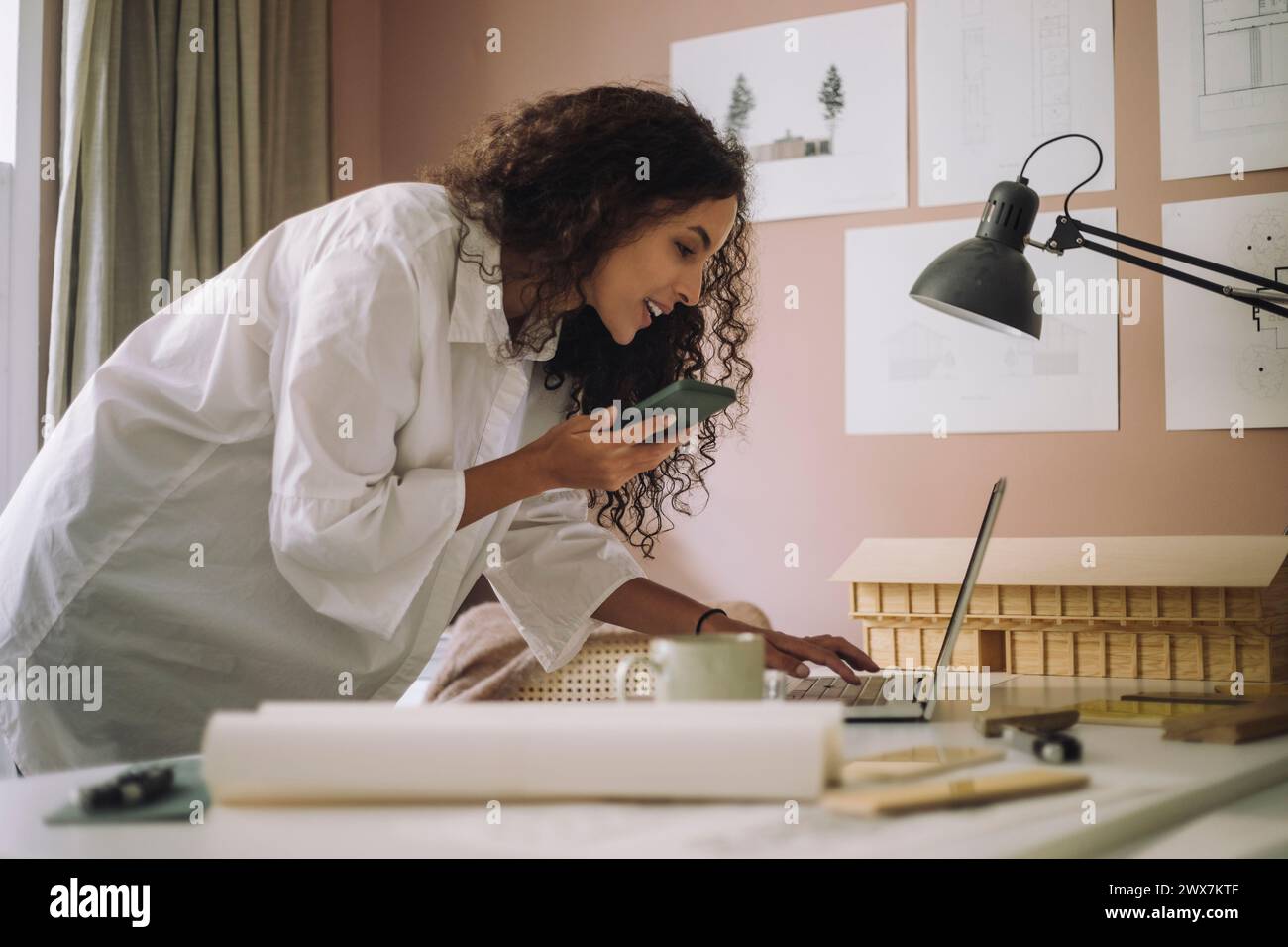 Smiling female construction freelancer talking through speaker phone and using laptop while working at office Stock Photo