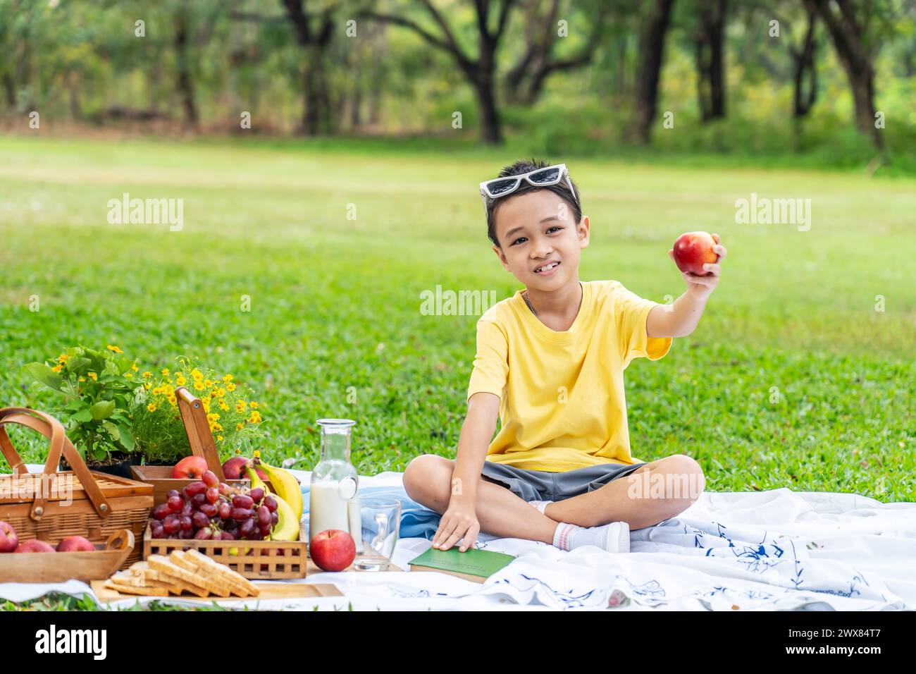 A child sitting on green grass, holding an apple, next to a picnic basket Stock Photo
