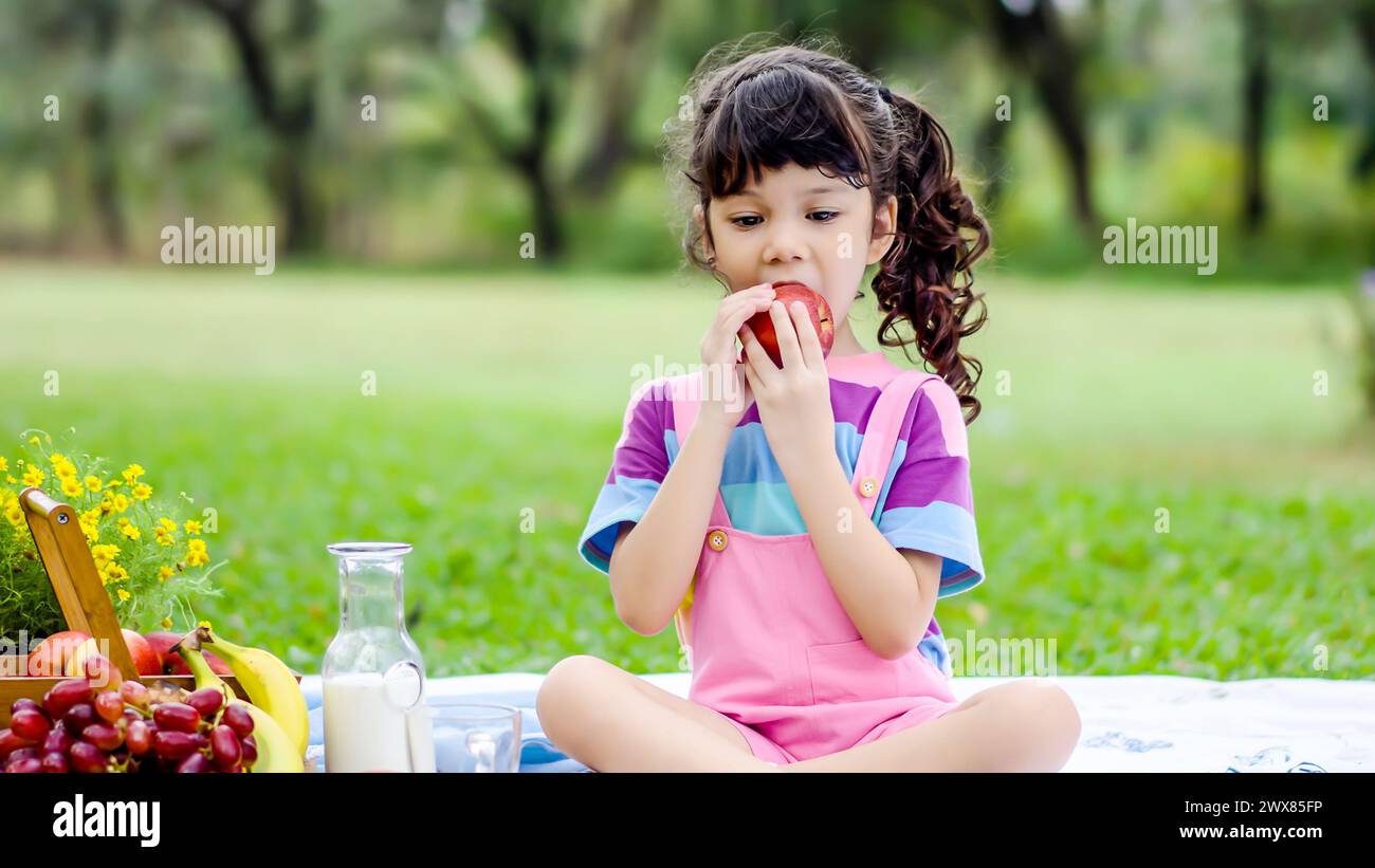 A young girl sits on grass, eating fruit and drinking milk Stock Photo