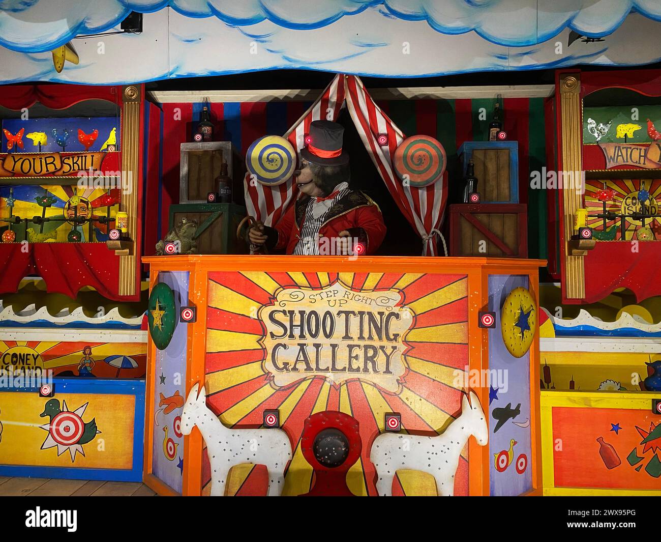 A vibrant vintage-style shooting gallery at a carnival with an animatronic bear dressed as a carnival barker inviting participants to test their aim Stock Photo