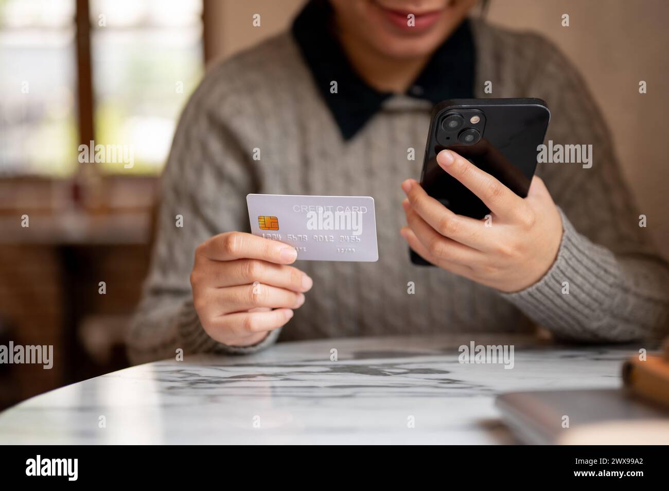 A cropped shot of an Asian woman holding her smartphone and a credit card, sitting at a table indoors, shopping online, registering a credit card acco Stock Photo