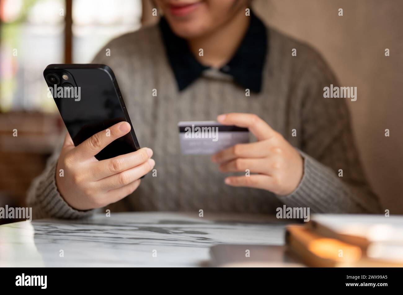 A cropped shot of an Asian woman holding her smartphone and a credit card, sitting at a table indoors, shopping online, registering a credit card acco Stock Photo