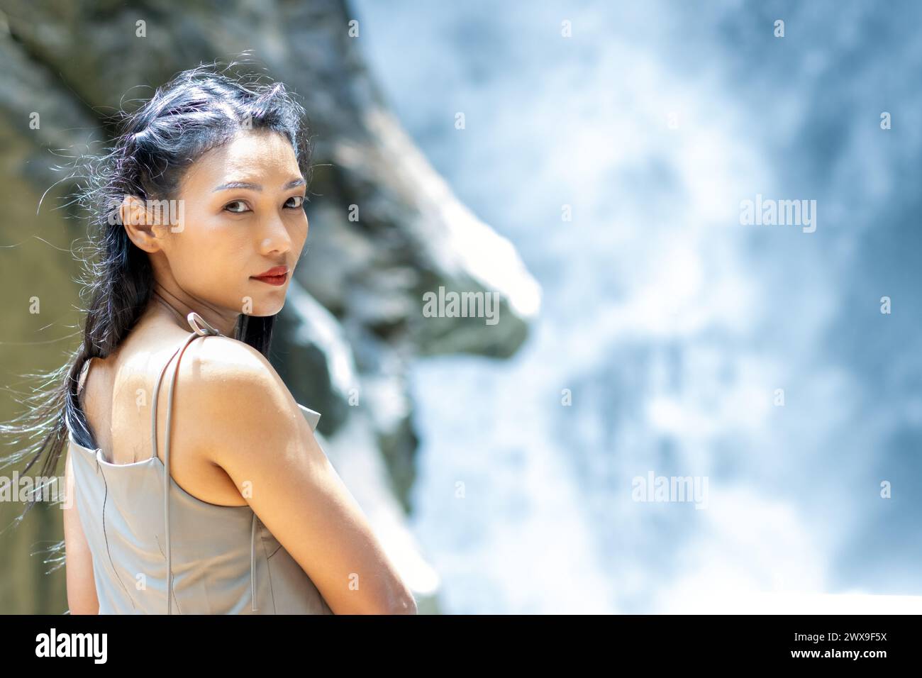 A young woman stands in front of a waterfall and turns to the camera ...
