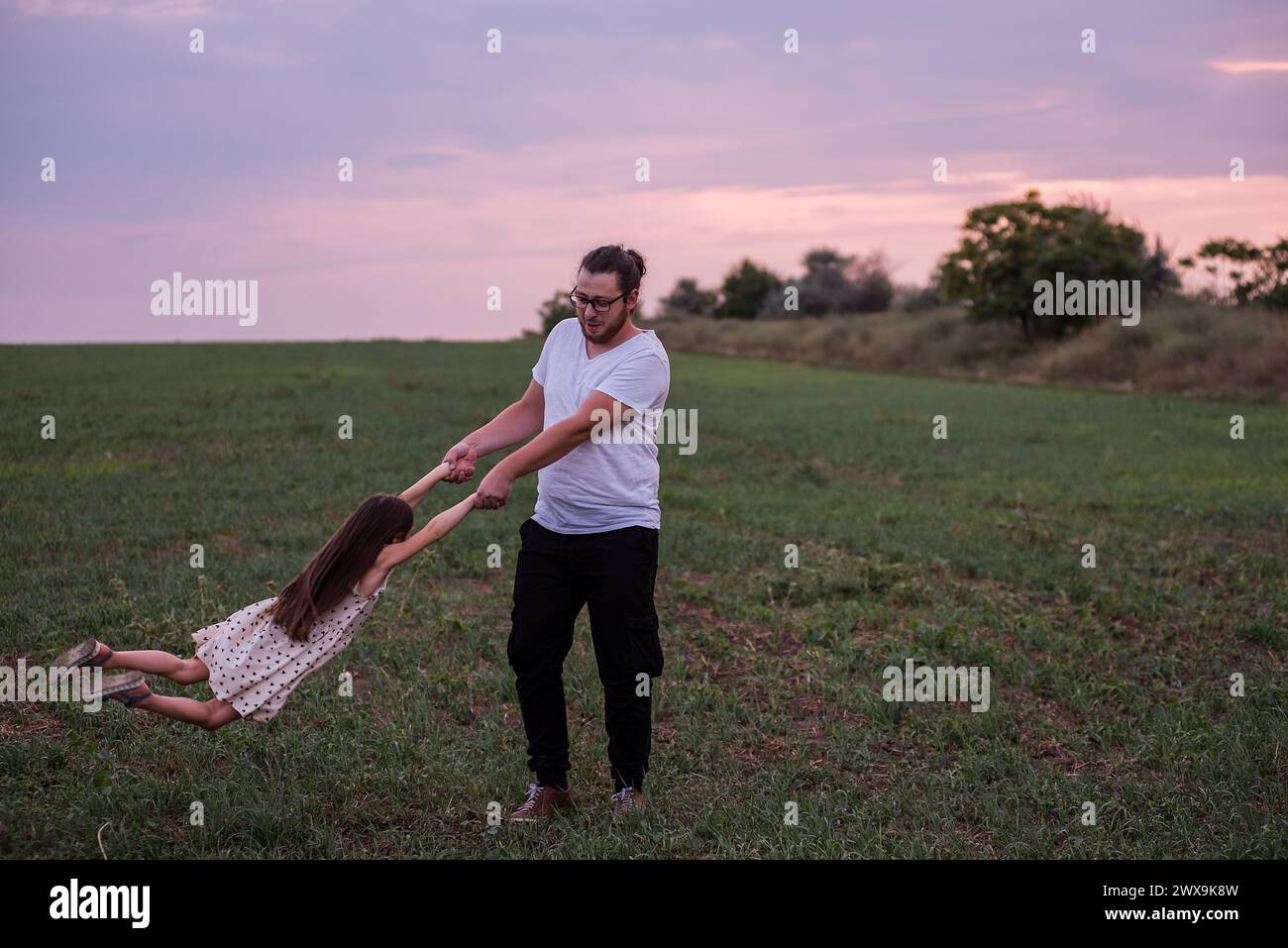 Young man and small girl enjoy playful moment, spinning around in grassy field under pastel sunset sky, conveying warmth, family fun. Joyful Father pl Stock Photo
