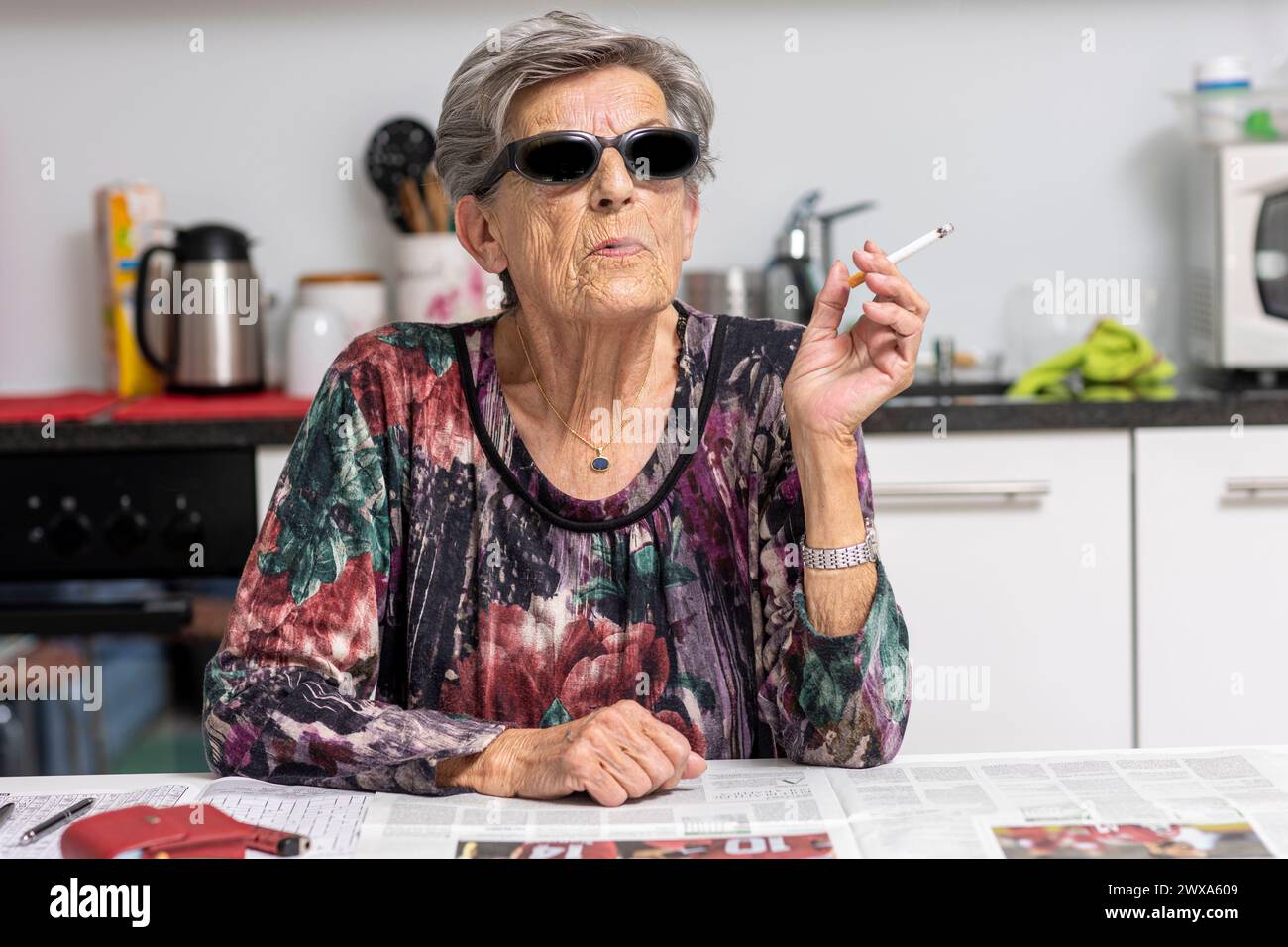 Portrait of old smoker sitting in her modern white kitchen with dark sunglasses. A woman from another time. Stock Photo