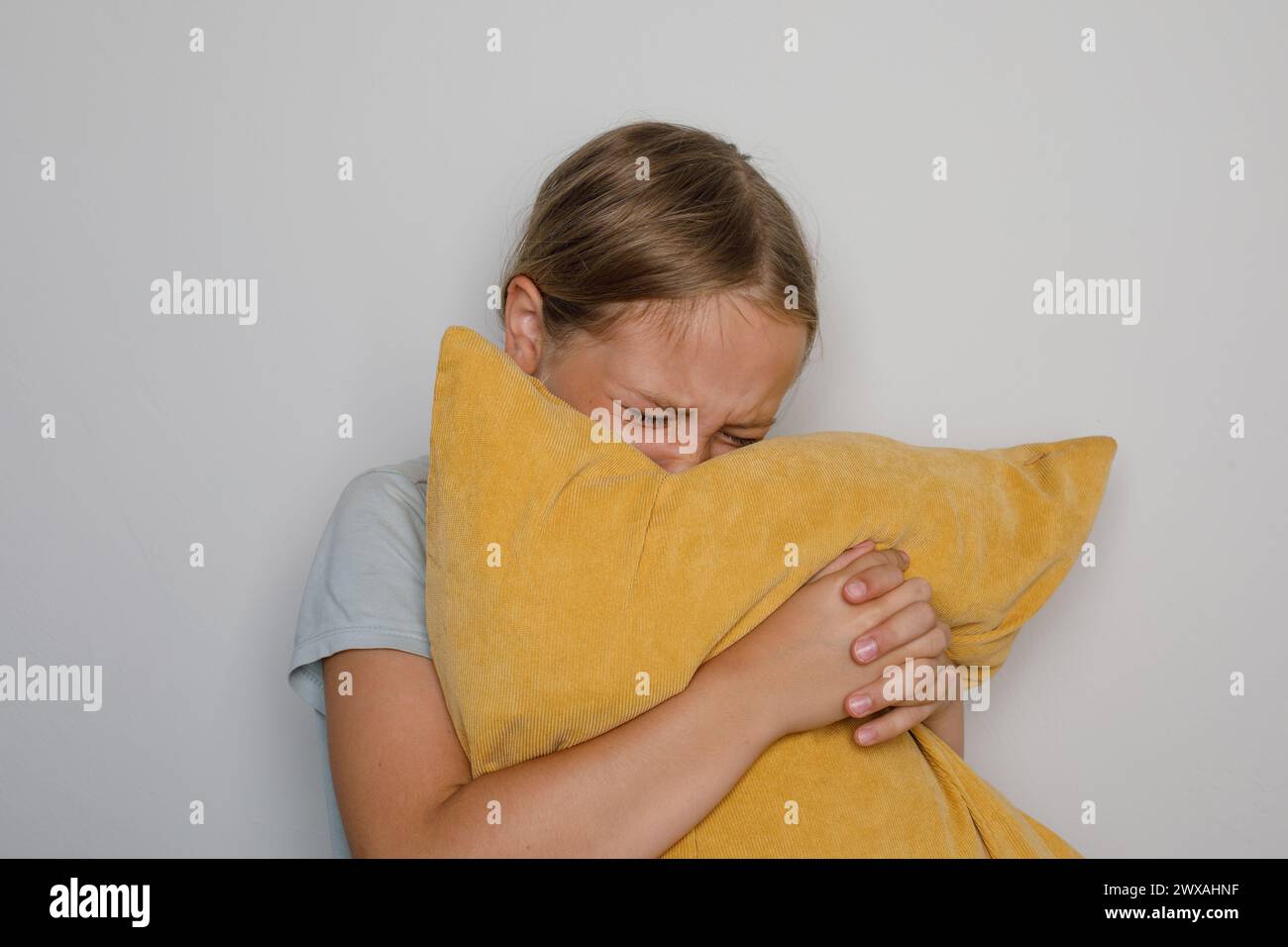 Cute child girl crying and embracing yellow pillow against white domestic wall at home Stock Photo