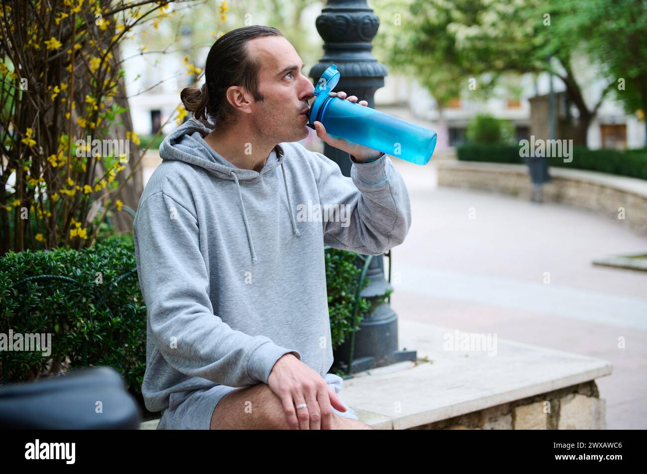 Caucasian young man in gray sportswear, drinking water from a sports bottle in the city Stock Photo