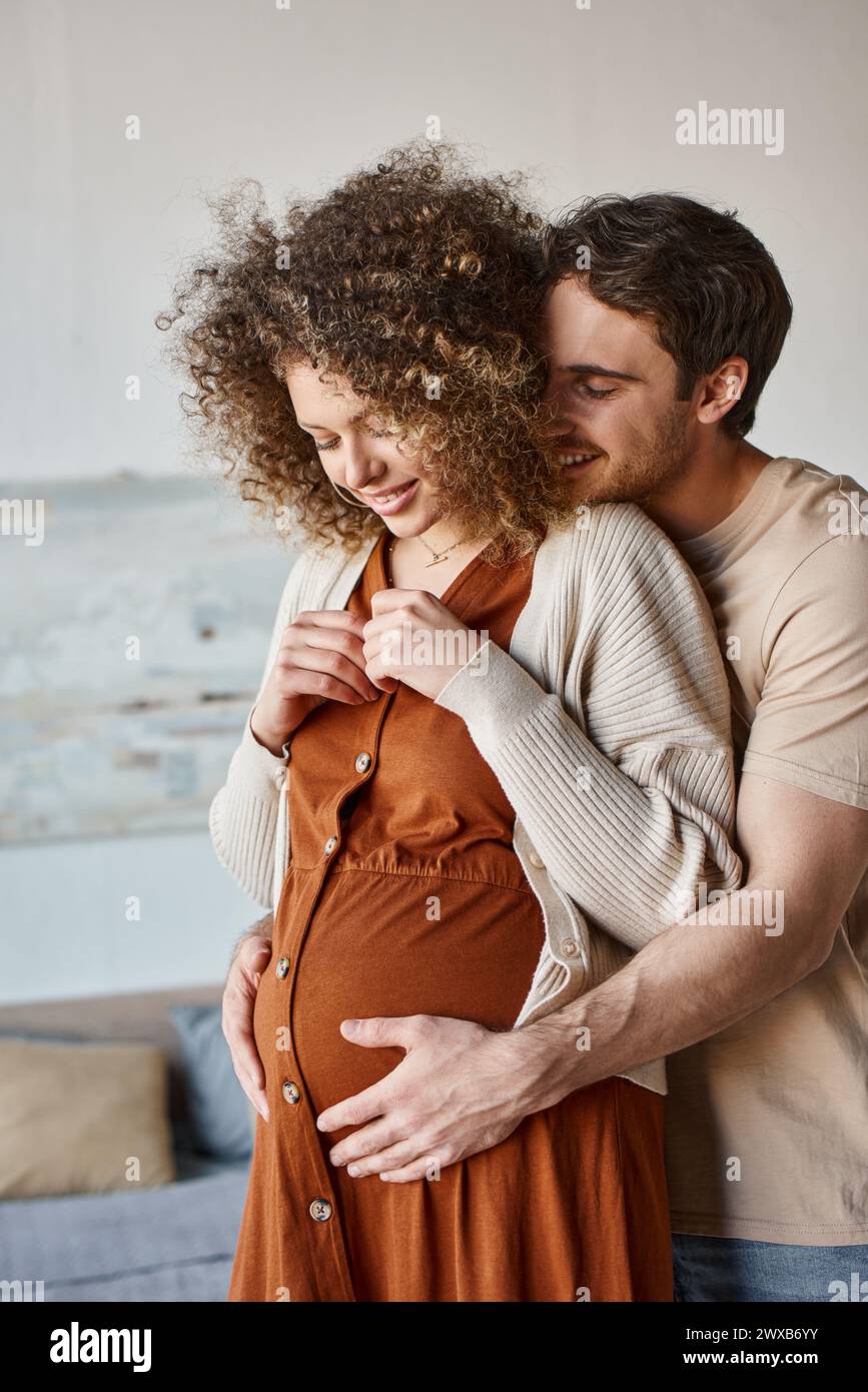 Lovely happy man hugging his pregnant wife from behind standing at home ...