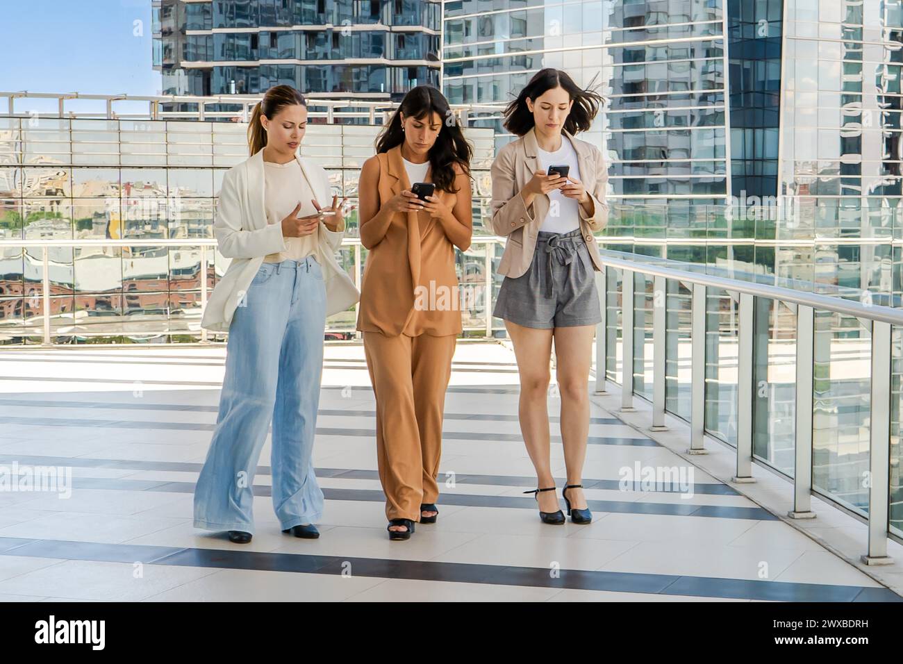 Three businesswomen are walking while using smartphones outdoors. All women are dressed in business attire Stock Photo