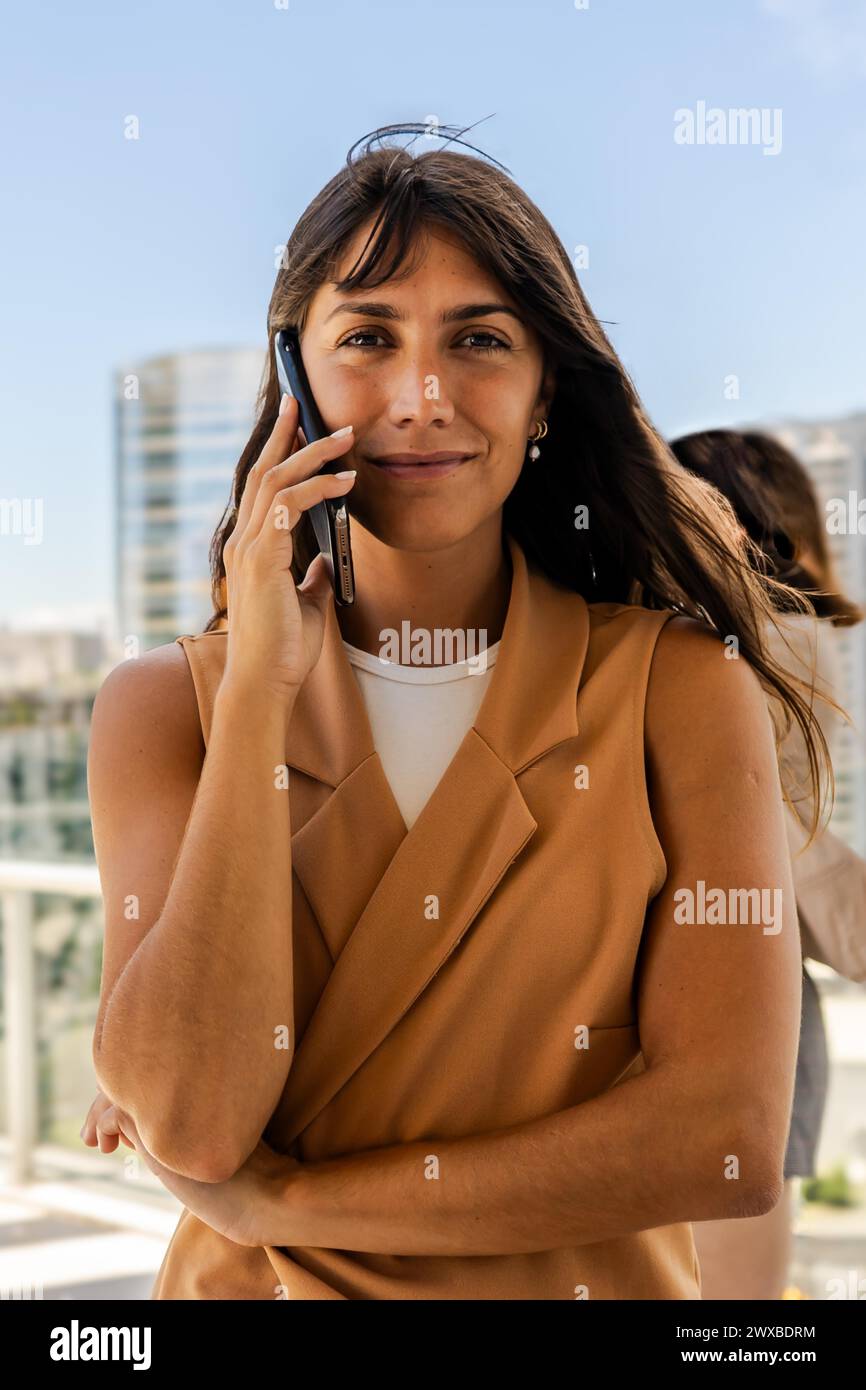 A businesswoman wearing a brown jacket smiles while talking on her cell phone and looking at the camera Stock Photo