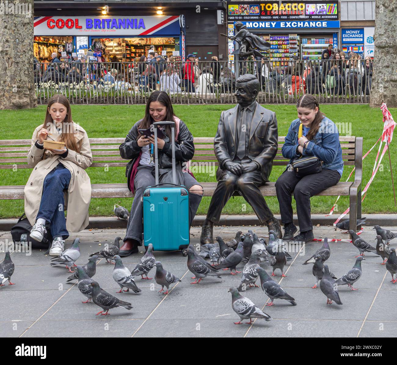 Women sitting on a bench next to a bronze statue of Mr Bean, in Leicester Square, London. Pigeons gather in front of them in search of food. Stock Photo