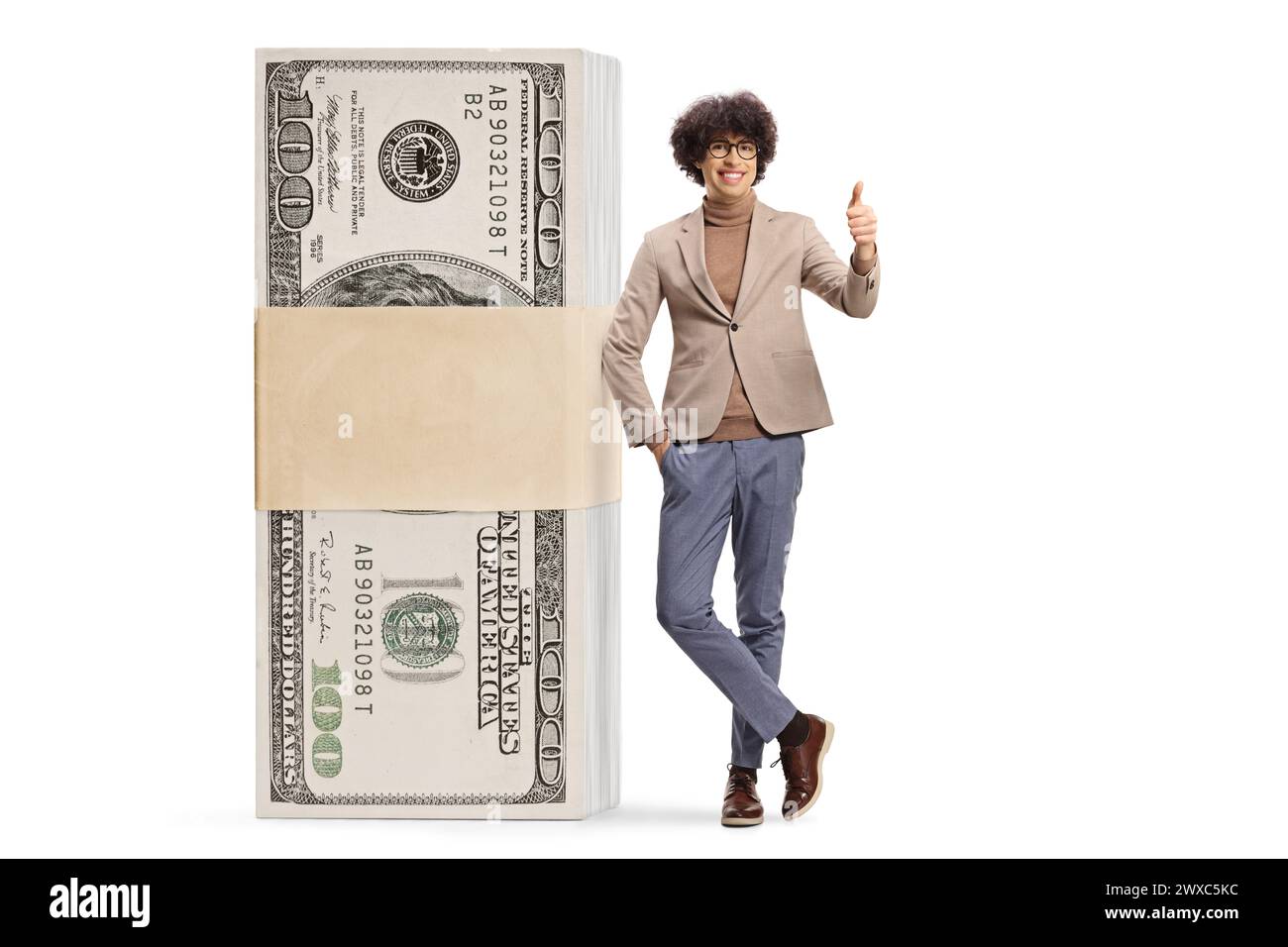 Young man leaning on a stack of money and gesturing thumbs up isolated on white background Stock Photo