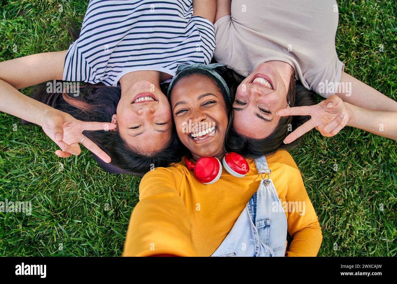 Selfie from above of three cheerful multiracial young women lying on grass outdoors. Gen z in park. Stock Photo