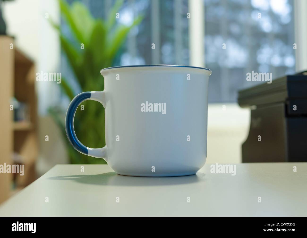 A enamel white mug with a blue handle, a type of drinkware, is placed on a white table, a piece of tableware or serveware Stock Photo