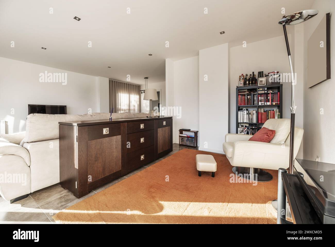 Living room of a house furnished with a white swivel armchair, a dark wood sideboard and a complete bookcase Stock Photo