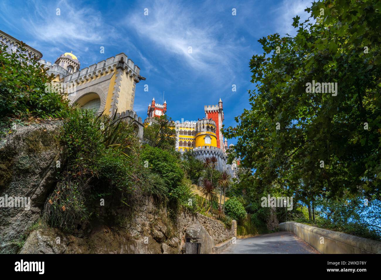 Exterior of the Pena Palace in a colorful yellow and red style on fortified walls covered with vegetation and palm trees under a sunny blue sky. Sintr Stock Photo