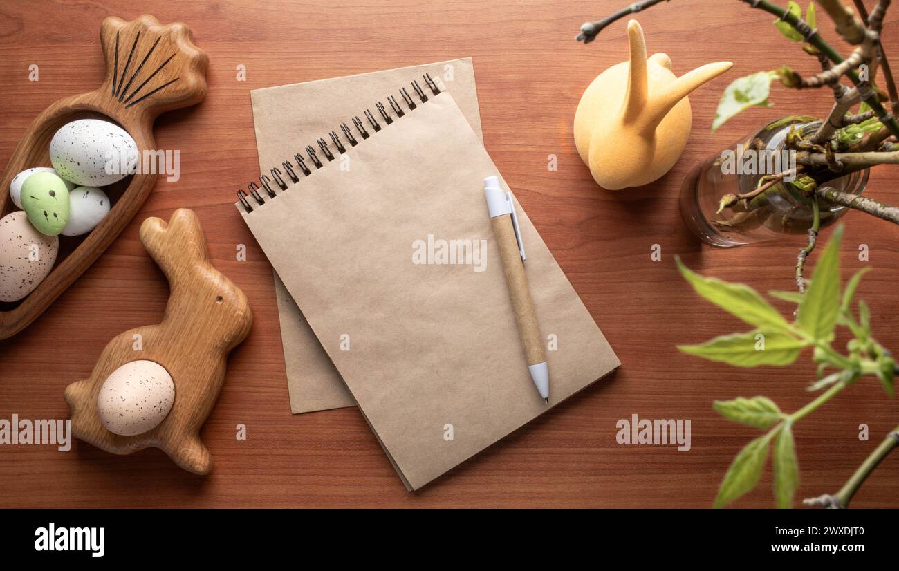 Blank craft paper notepad, pen among Easter decorations figured wooden plates in form bunny and carrot with eggs, twigs with spring leaves on wooden d Stock Photo