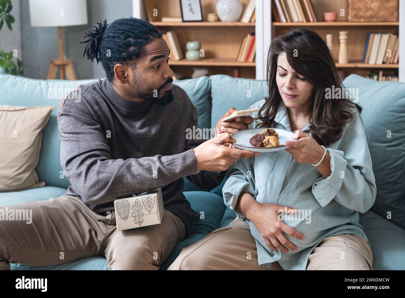 Pregnancy support and fun. Young couple man and pregnant woman sitting at home teasing with sweet food forbidden by doctor. Male and female support an Stock Photo