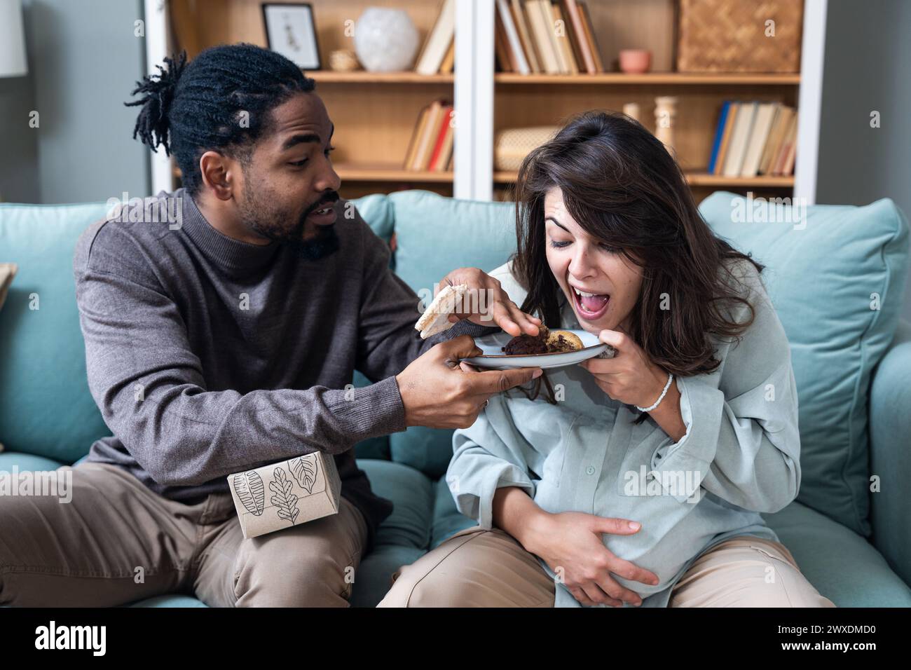 Pregnancy support and fun. Young couple man and pregnant woman sitting at home teasing with sweet food forbidden by doctor. Male and female support an Stock Photo