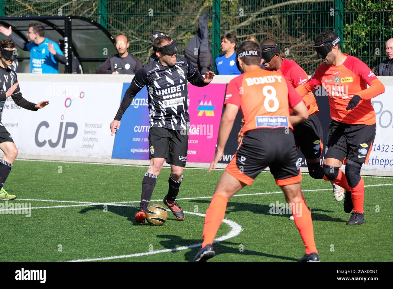 Royal National College for the Blind, Hereford, UK – Saturday 30th March 2024 – Round 3 of the European Blind Football League ( EBFL ) held at the Royal National College for the Blind at Hereford. The ball is fitted with ball bearings that rattle to assist the players to locate it. A player from Belgian team Cecifoot Charleroi is surrounded by three players from French club Bondy Cecifoot Club ( orange shirts ).  Photo Steven May / Alamy Live News Stock Photo