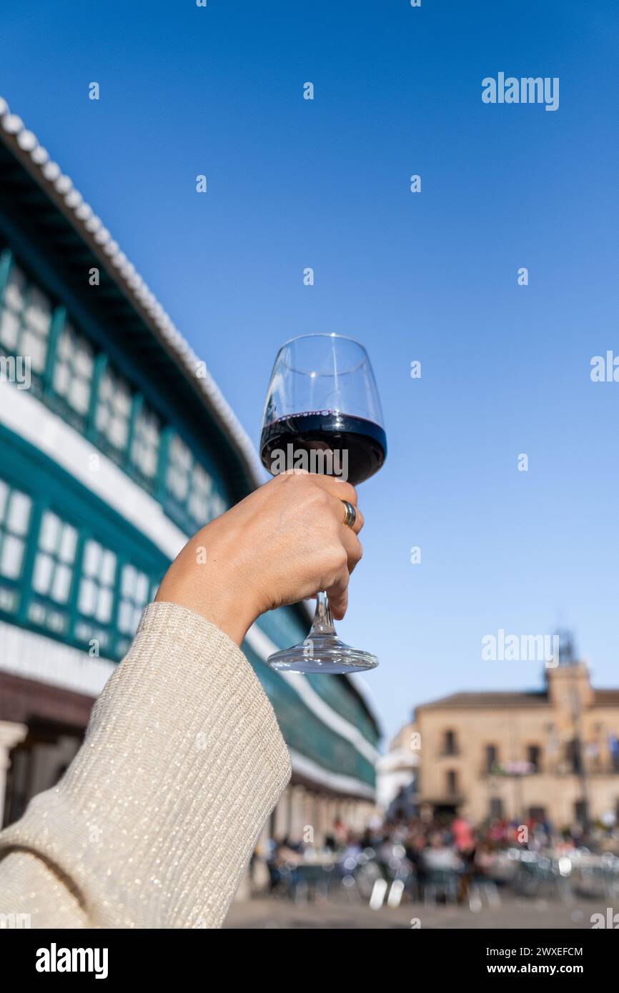 An elegant woman's hand holds a glass of wine with the main square of Almagro in the background out of focus on a sunny day. It is a local wine highly Stock Photo