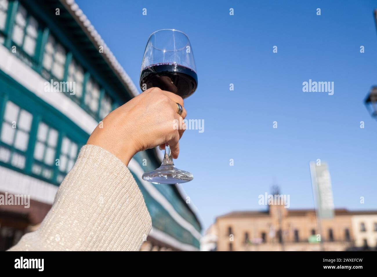 A woman's hand holds a glass of wine with the main square of Almagro in the background out of focus on a sunny day. It is a local wine highly apprecia Stock Photo