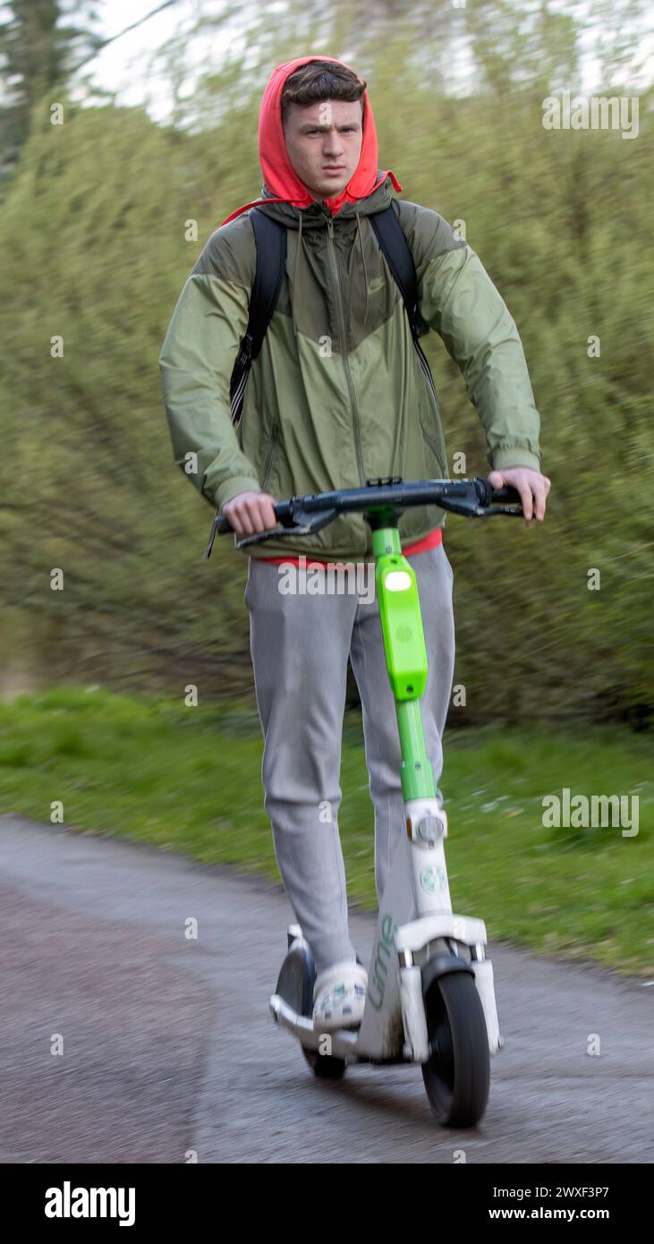 Milton Keynes,UK-Mar 30th 2024:  Young Man Riding an Electric Scooter, set against a backdrop of greenery Stock Photo