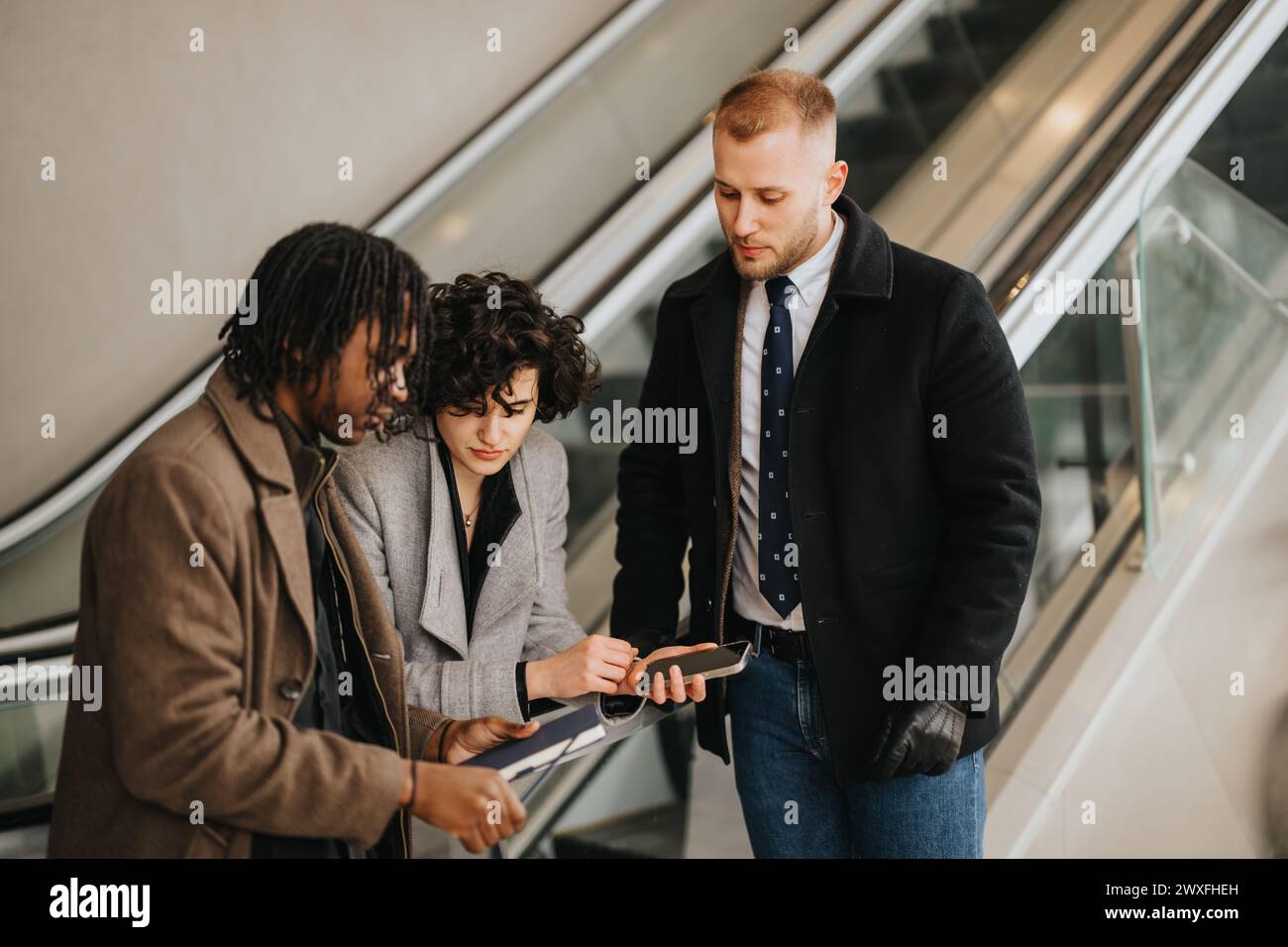 Three people colleagues in smart attire discussing over a tablet while standing on an escalator in a corporate setting. Stock Photo