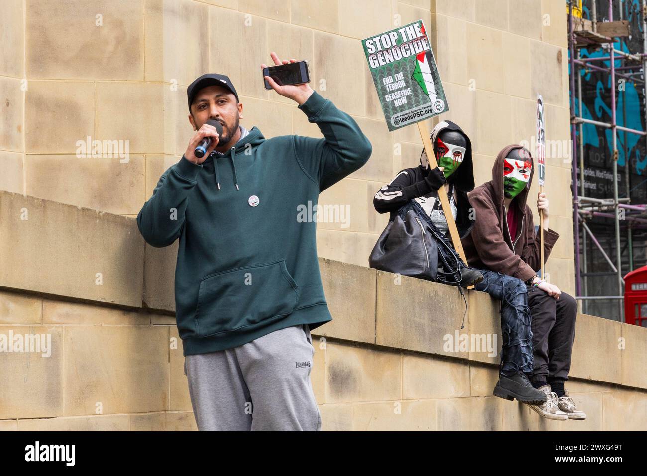 Leeds, UK. 30 MAR, 2024. Pro Palestine protest organiser speaks at Leeds PSC event with 2 masked demonstrators holding placards watching on in the background. Following speeches protestors marched through the city center of Leeds. Credit Milo Chandler/Alamy Live News Stock Photo