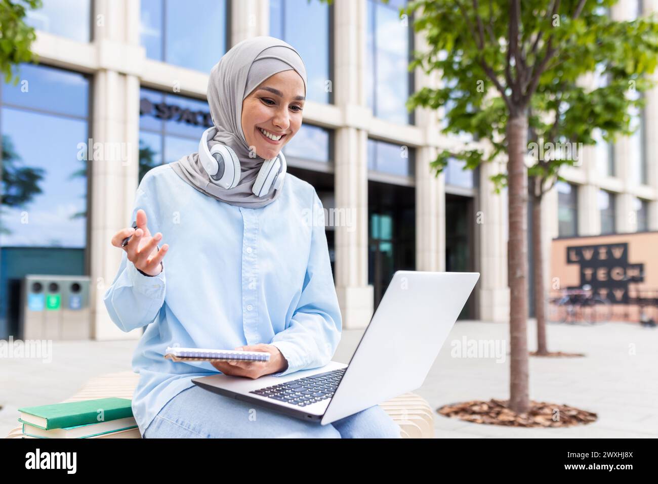 Focused young student wearing a hijab with headphones studying on her laptop and taking notes outside on campus. Stock Photo