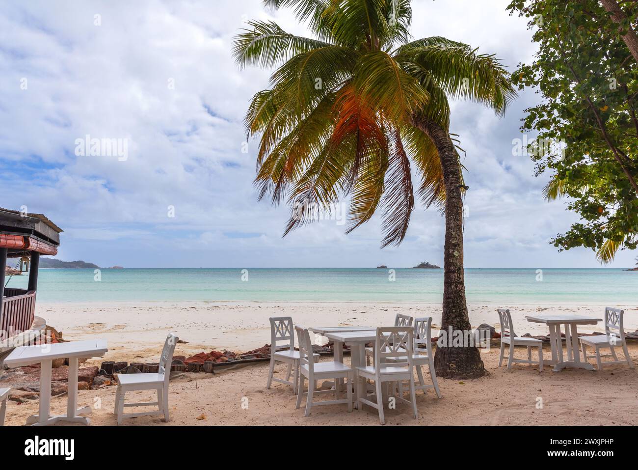 Seychelles seaside view with white wooden tables and chairs under coconut palm trees. Cote D'Or Beach, Praslin island Stock Photo
