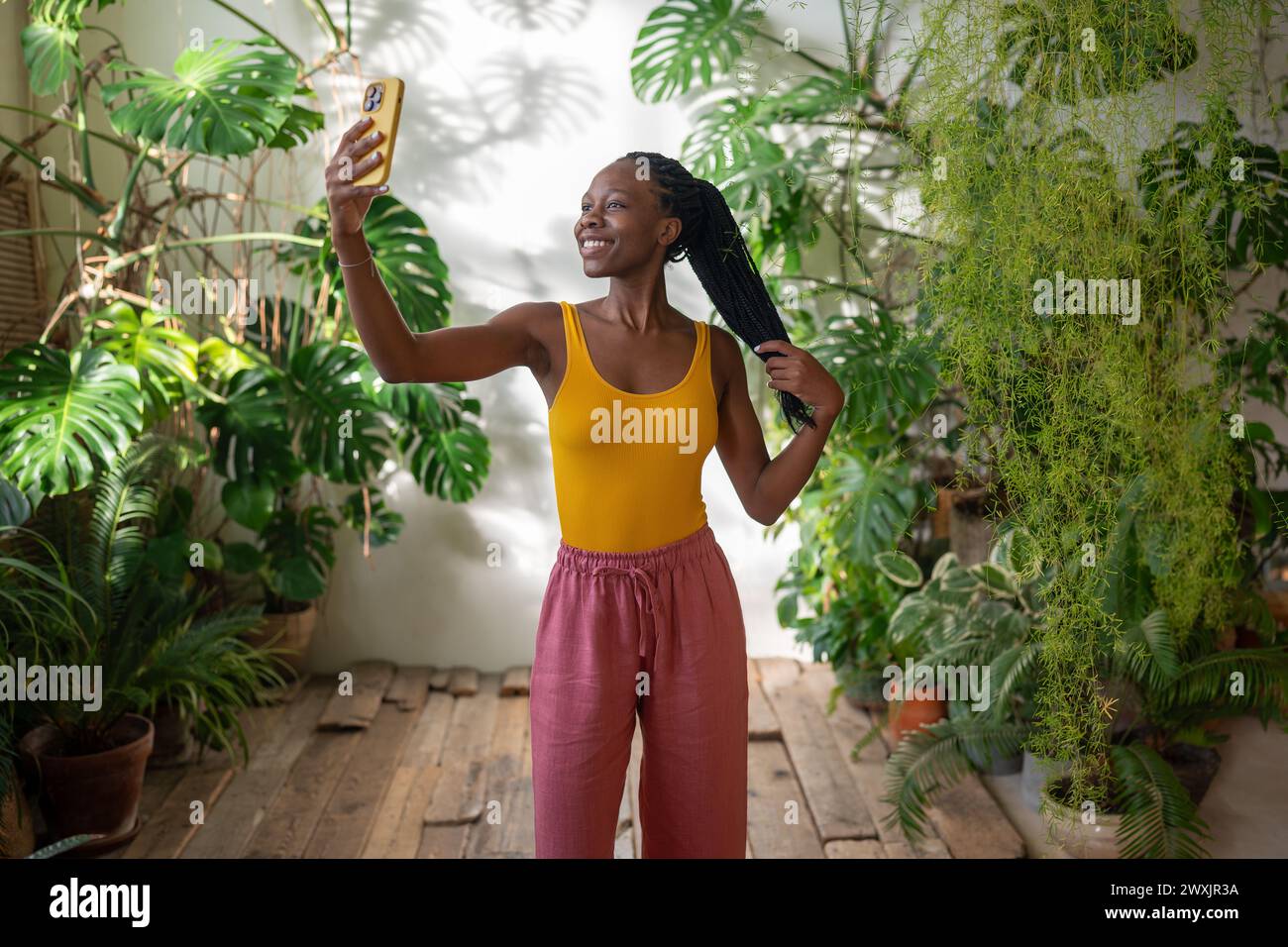 Happy joyful black woman smiling makes selfie on smartphone stands in room with tropical houseplants Stock Photo