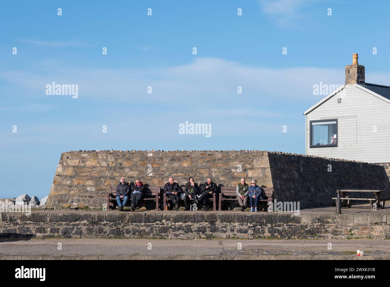 31 March 2024. Cullen,Moray,Scotland. This is a gathering of people enjoying the sunshine and seated on benches at Cullen Harbour. Soaking up the suns Stock Photo