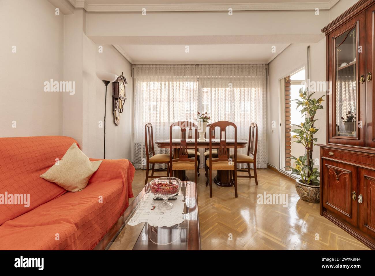 living room with reddish wood table with matching glass bookcase, fabric upholstered sofa and dining table next to a large window with curtains and ac Stock Photo