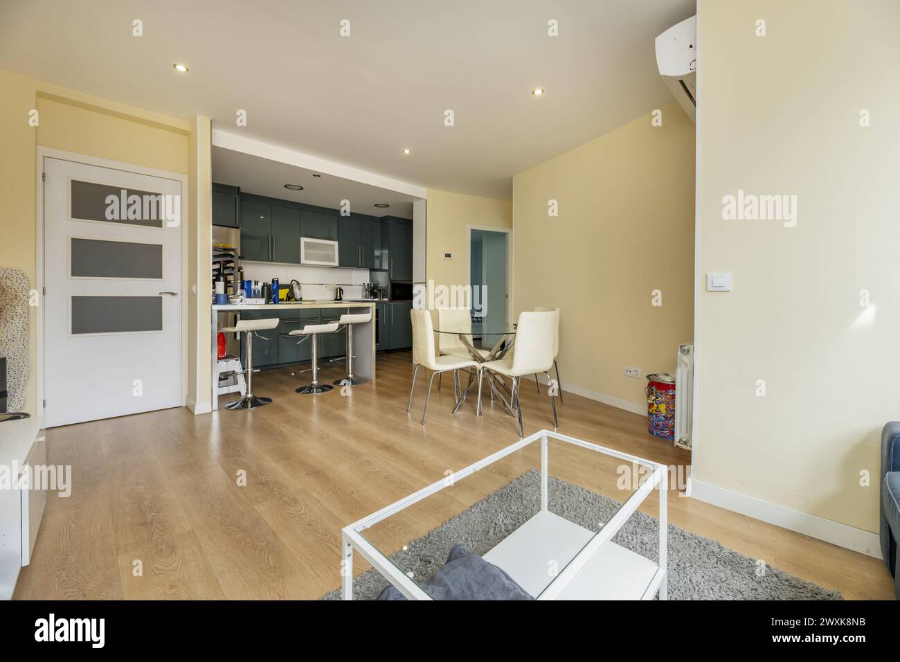 Living room with round glass table with white chairs, white door, open kitchen and a kitchen island with high chrome stools Stock Photo