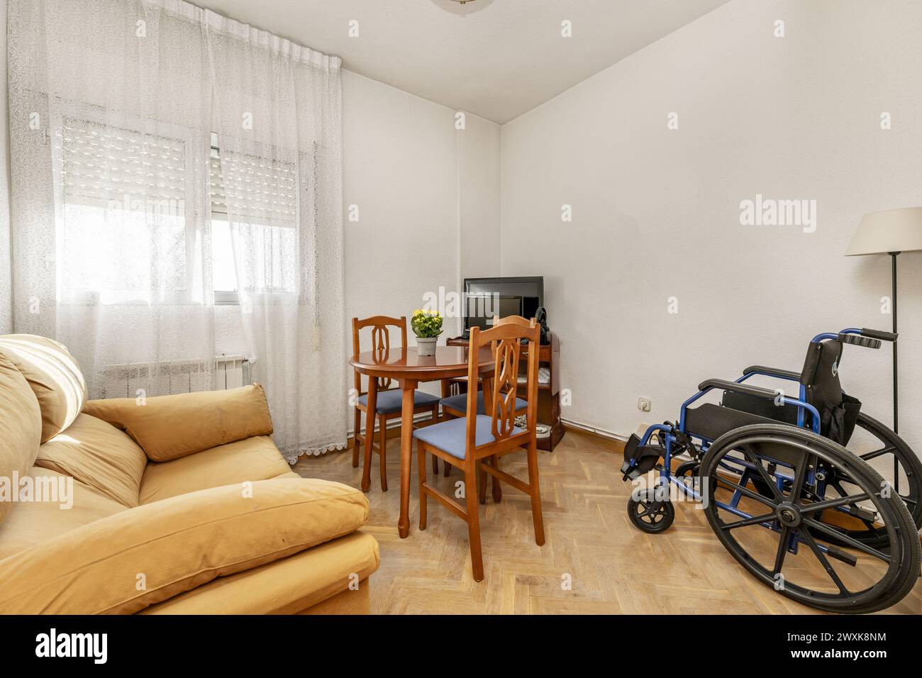 Corner of a small living room with round wooden table with matching chairs upholstered in blue microfiber with a black wheelchair next to it Stock Photo