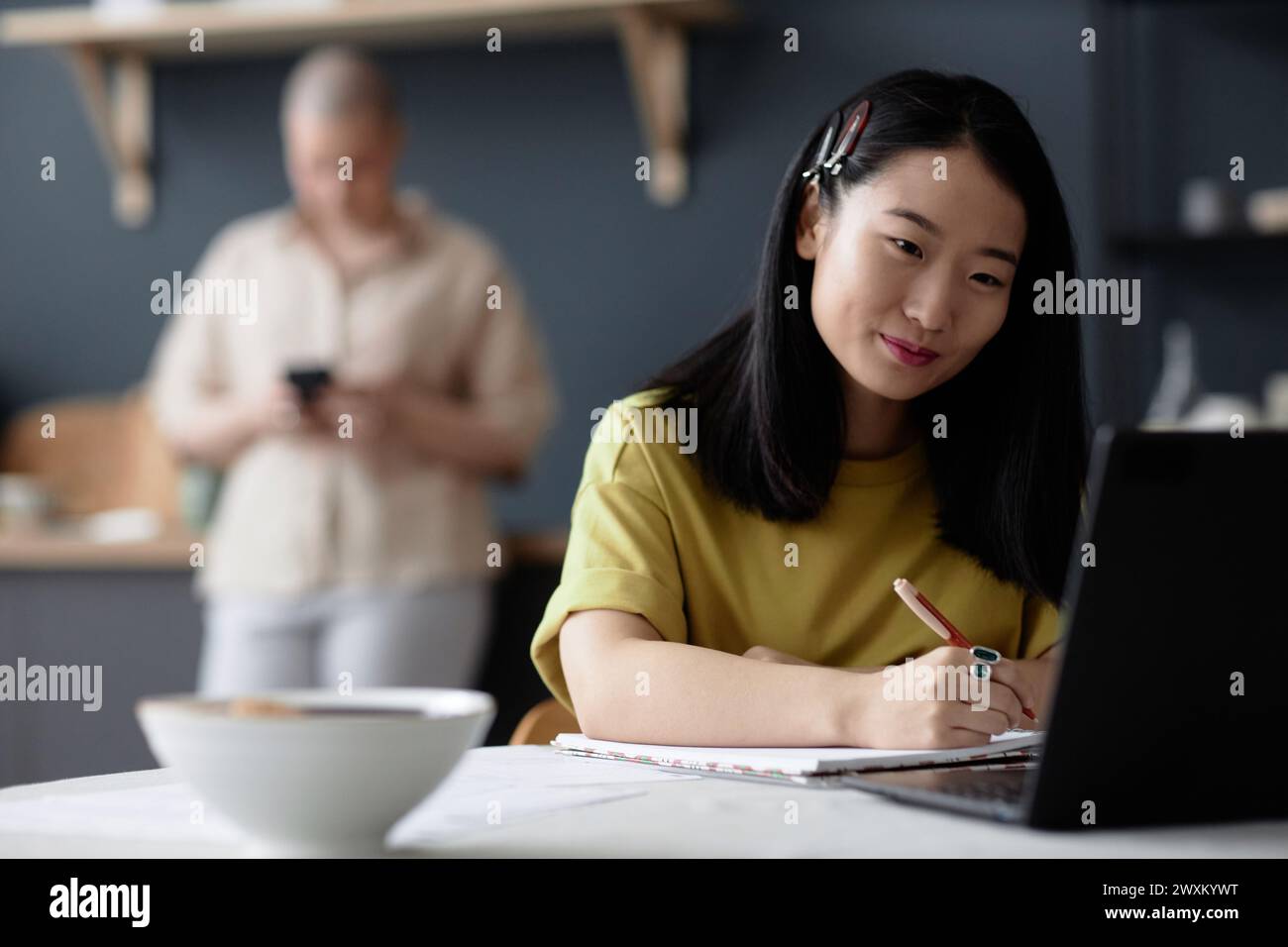 Young female Asian student sitting at dining table looking at laptop ...
