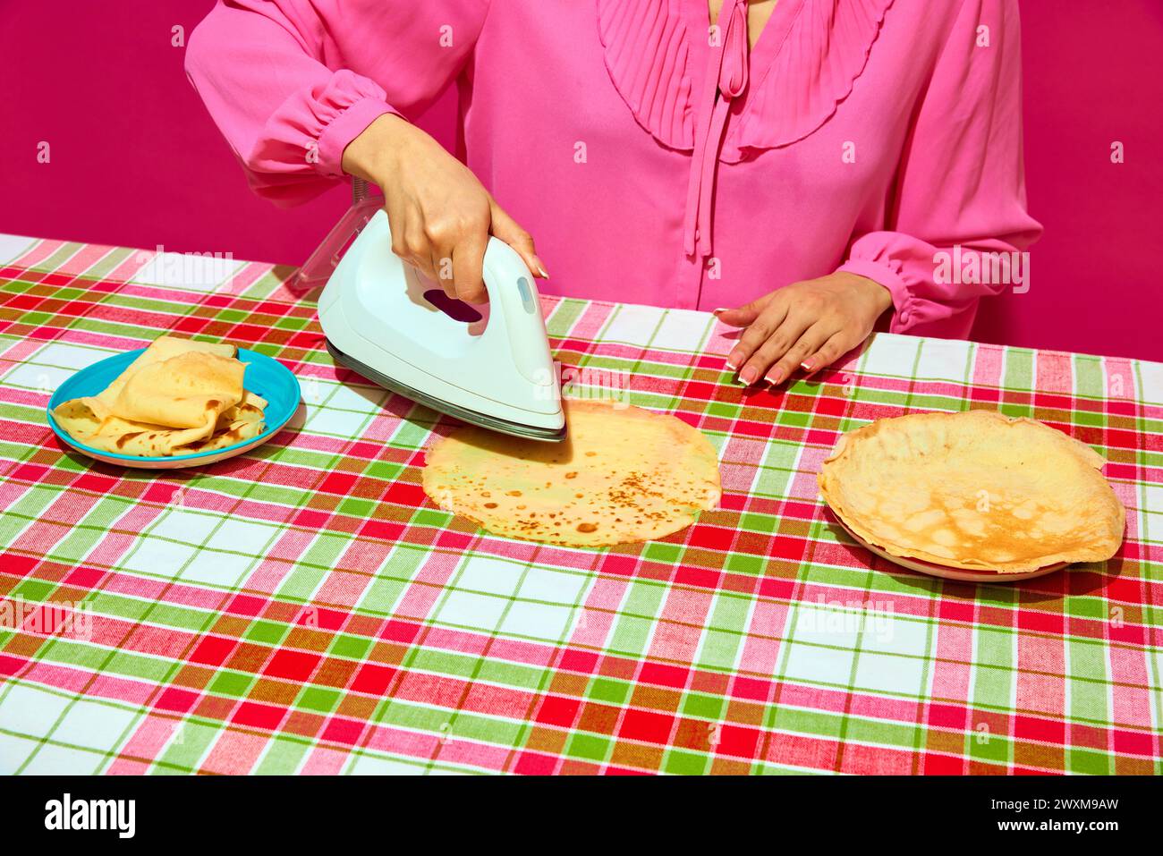 Woman in pink blouse ironing pancake on plaid tablecloth. Meme featuring unexpected ways to prepare food through humor. Stock Photo